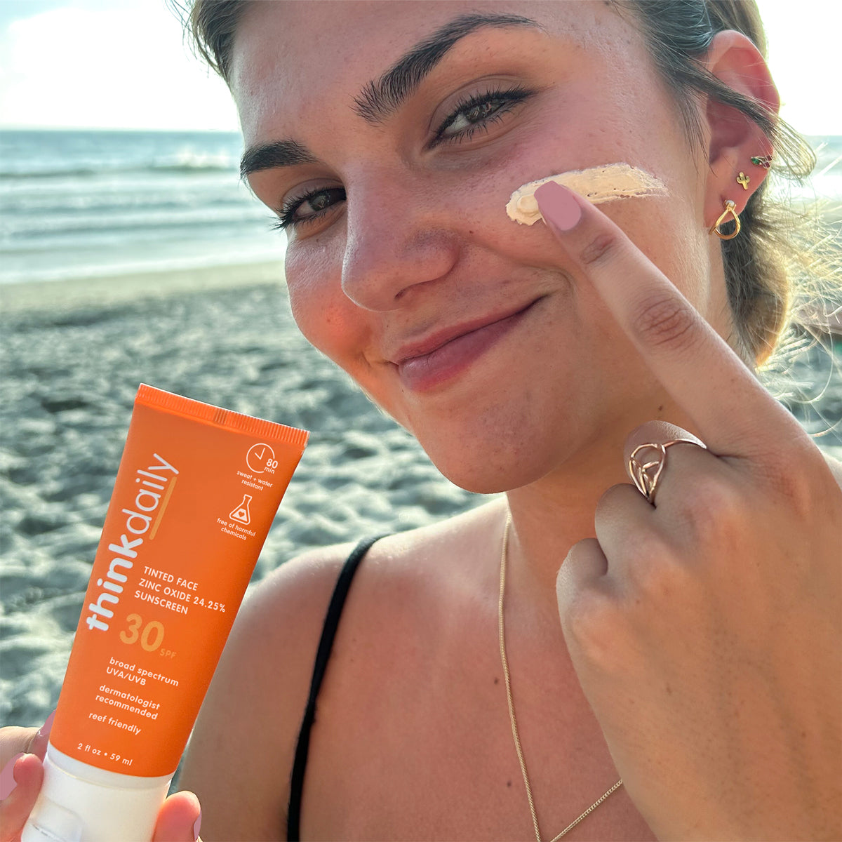 Woman applying ThinkDaily Tinted Face 30 SPF Sunscreen on the beach, emphasizing broad-spectrum UV protection, dermatologist recommended, and reef-friendly formula.