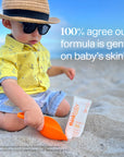 A toddler on a beach, wearing a hat and sunglasses, sitting next to a Thinkbaby sunscreen tube with text stating "100% agree it's gentle on baby’s skin".