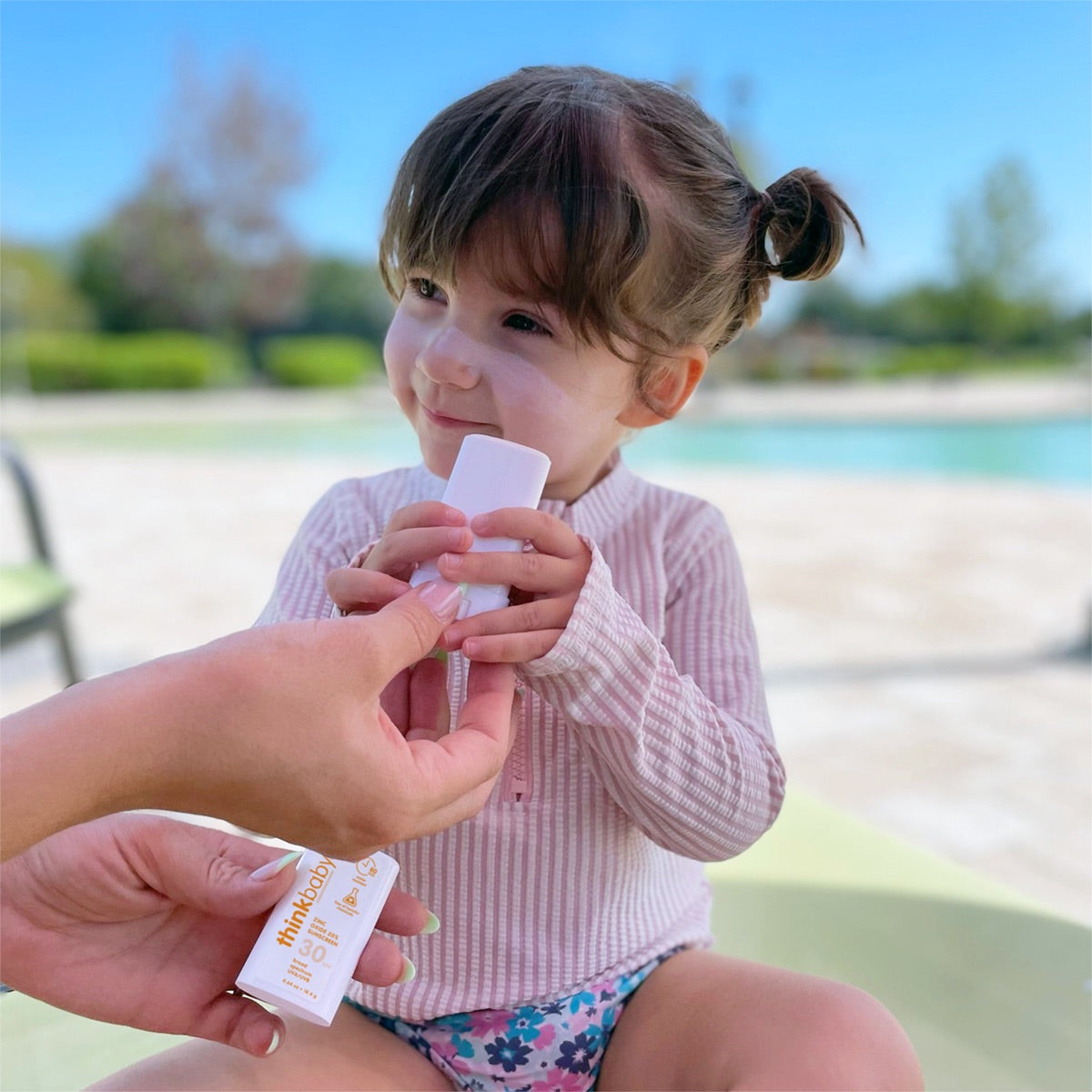 A young child smiling while applying Thinkbaby sunscreen stick with an adult&#39;s hand holding the tube, by a poolside on a sunny day.