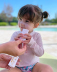A young child smiling while applying Thinkbaby sunscreen stick with an adult's hand holding the tube, by a poolside on a sunny day.