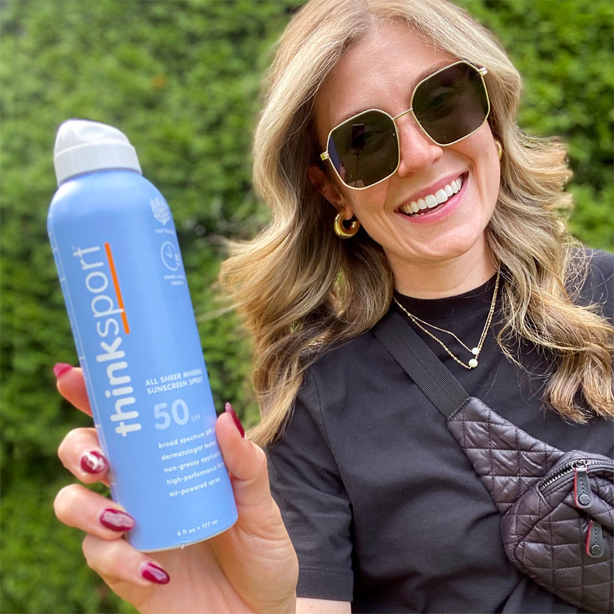 A smiling woman holding a ThinkSport sunscreen spray with SPF 50, showcasing the product outdoors with a natural backdrop.