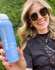 A smiling woman holding a ThinkSport sunscreen spray with SPF 50, showcasing the product outdoors with a natural backdrop.